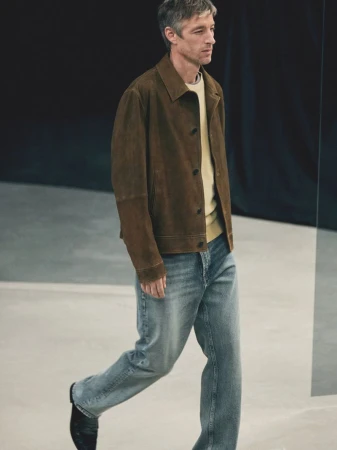 An adult man in a brown suede jacket and jeans walks in a photo studio with a black backdrop and concrete floor, shot in medium perspective.