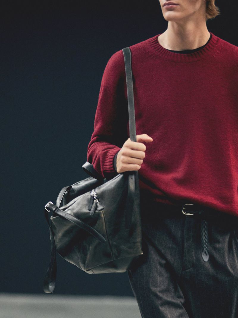 Man wearing a red sweater and carrying a black leather duffel bag, showcasing fashion and accessories against a dark background.
