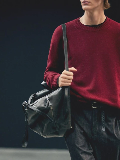 Man wearing a red sweater and carrying a black leather duffel bag, showcasing fashion and accessories against a dark background.