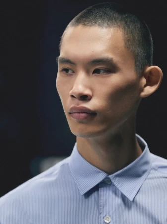 Close-up portrait of model Yura Nakano with a buzz cut wearing a light blue collared button-down shirt against a dark background.