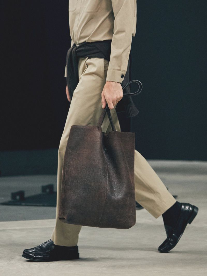 Model walking with a large brown leather tote bag, wearing beige trousers, a matching shirt, and polished black loafers.