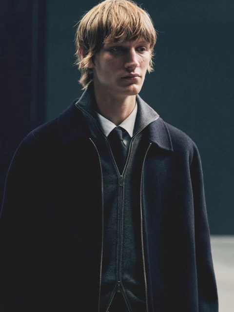 Close-up of a blonde male model in a navy coat, gray zip-up sweater, white shirt, and tie, posing indoors against a dark background.