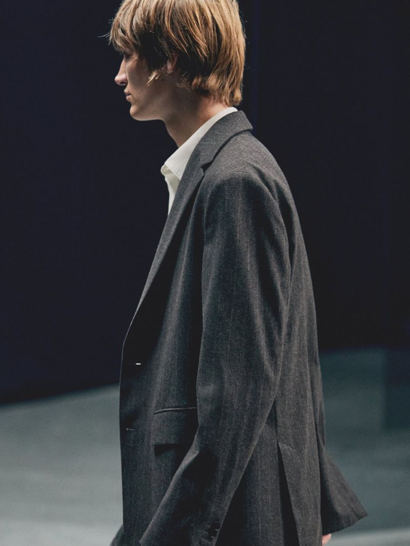 Profile of a man in a tailored suit and white shirt, captured in a photo studio setting with dark background and neutral concrete surface.