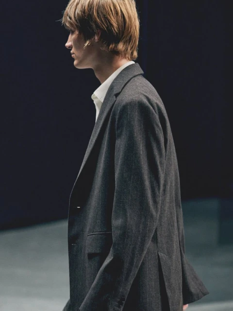 Profile of a man in a tailored suit and white shirt, captured in a photo studio setting with dark background and neutral concrete surface.
