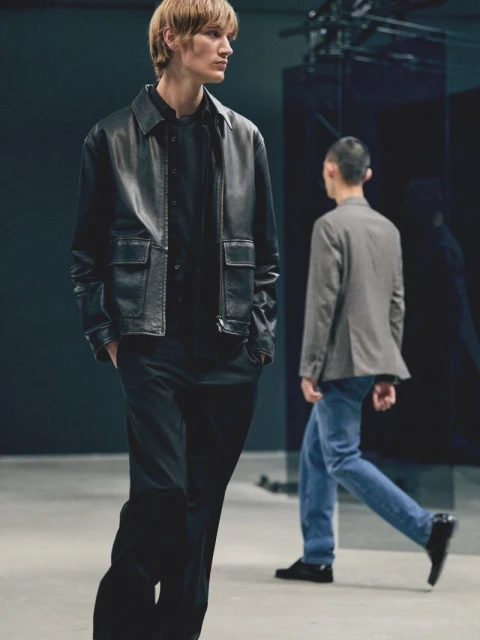 Two men are walking in an indoor fashion studio, one in a leather jacket and the other in a blazer and jeans, against a dark backdrop.