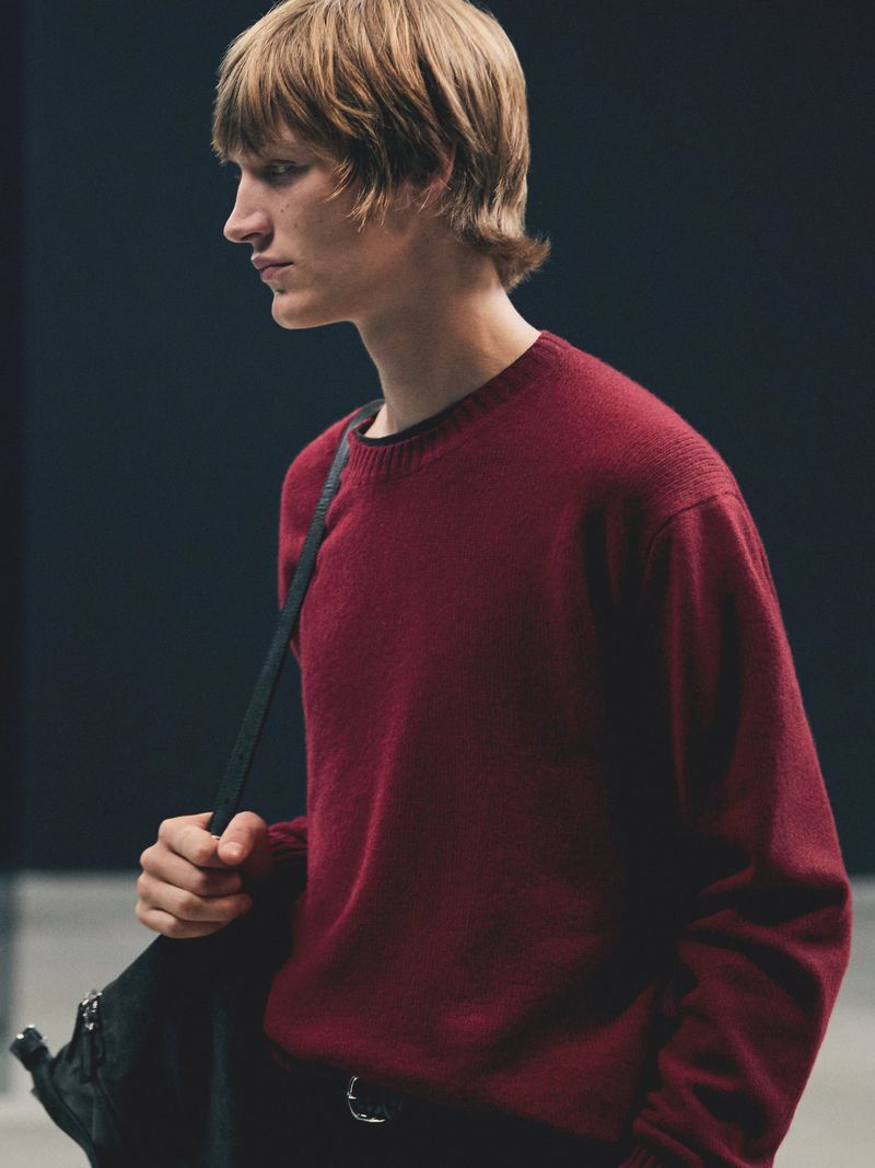 Headshot of a young adult male wearing a dark red crew neck sweater and carrying a black bag over his shoulder, against a dark background.