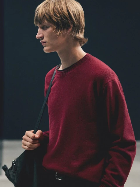Headshot of a young adult male wearing a dark red crew neck sweater and carrying a black bag over his shoulder, against a dark background.