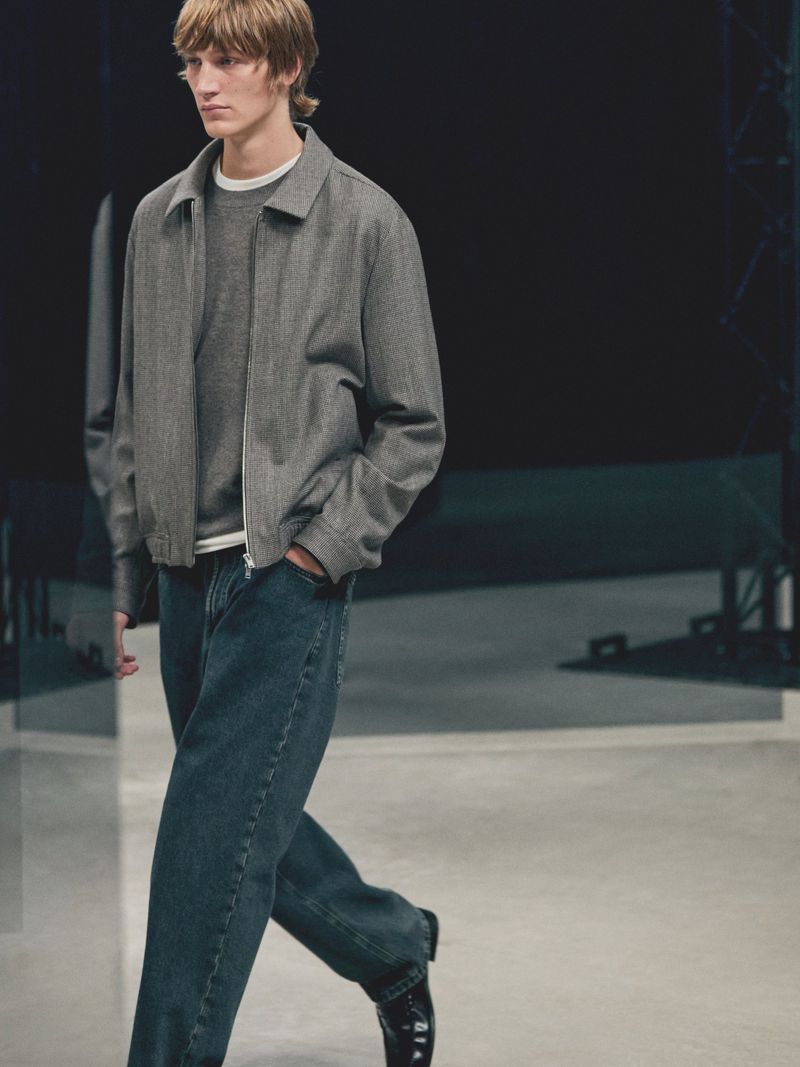 Model walking on a runway wearing a grey zip-up jacket, grey crewneck knit, and loose dark wash jeans for Massimo Dutti.