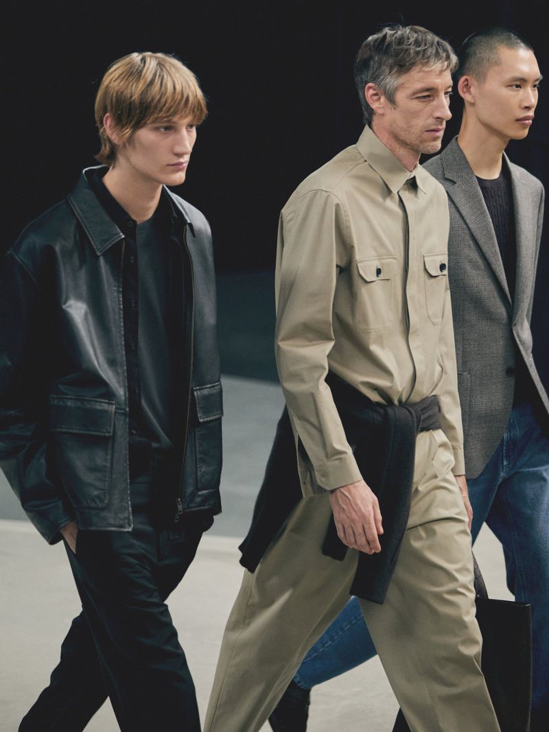 Three adult male fashion models walk in studio setting. One wears a leather jacket, another a tan button-up shirt, and another a blazer.