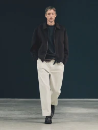 Male model walking in a dark zip jacket and wide-leg off-white trousers on a runway against a dark seamless backdrop.