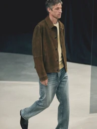 A male model walking on a runway wearing a brown suede jacket, yellow sweater, and blue denim jeans.
