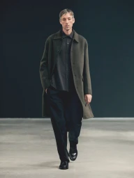 Male model walking forward wearing an olive green wool overcoat, dark collared shirt, black trousers, and black leather shoes.