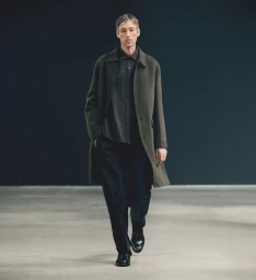 Model wearing an olive green wool coat, dark button-down shirt, and black trousers walking forward in a minimalist studio.