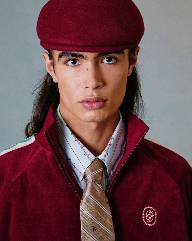Portrait of a young adult male with long dark hair wearing a burgundy cap, jacket, patterned shirt, and tie in a studio setting.