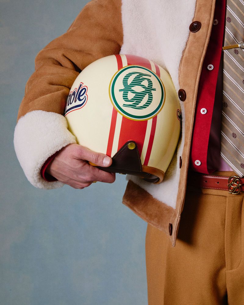 Person in a brown jacket holds a motorcycle helmet with racing stripes and logos, set against a simple blue background.