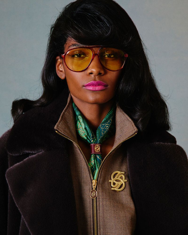A fashionable African American woman wearing sunglasses and a brown fur coat poses indoors, showcasing retro fashion with a green patterned scarf.