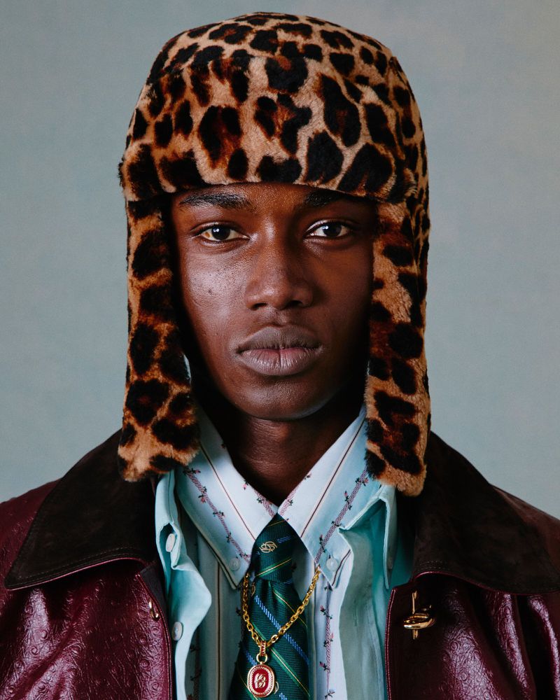 Close-up studio shot of a dark-skinned male model wearing a leopard print hat, patterned shirt, and patterned tie with a gold chain.