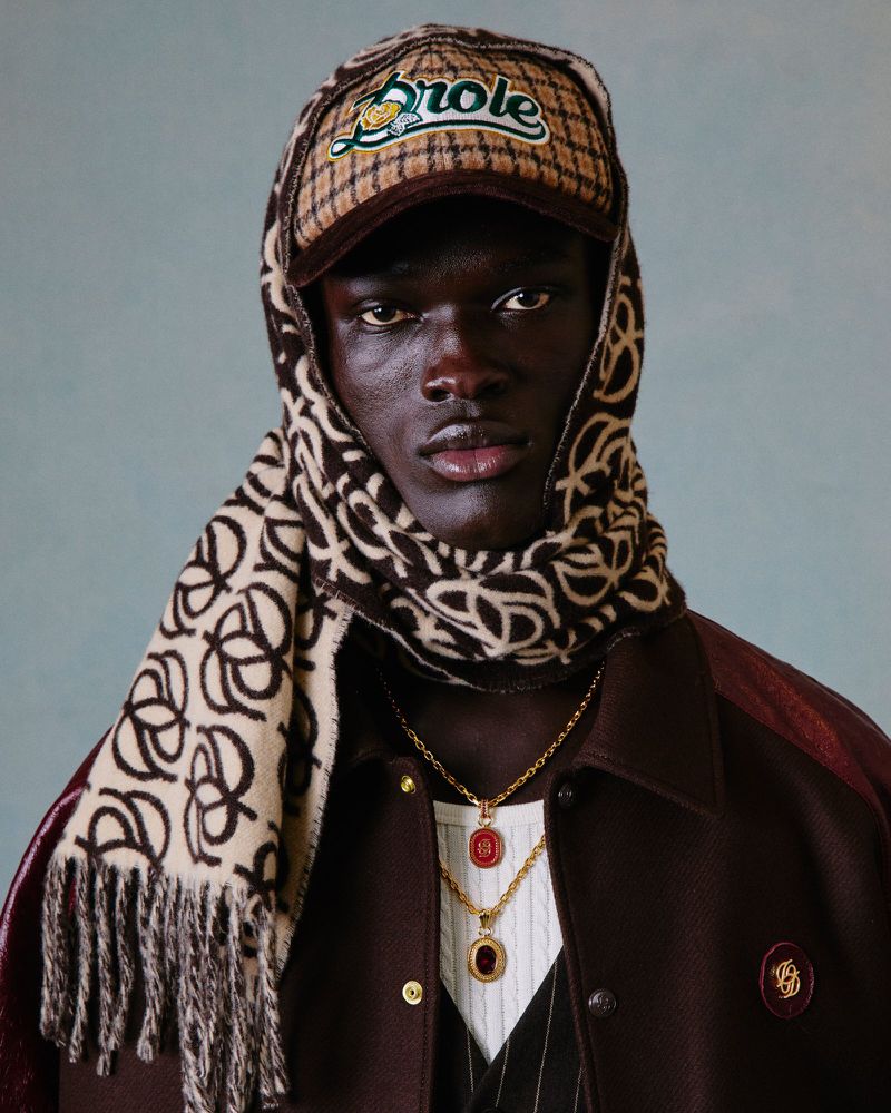 Portrait of a dark-skinned male wearing a patterned hat, scarf, gold chains, and a brown jacket against a light blue background.