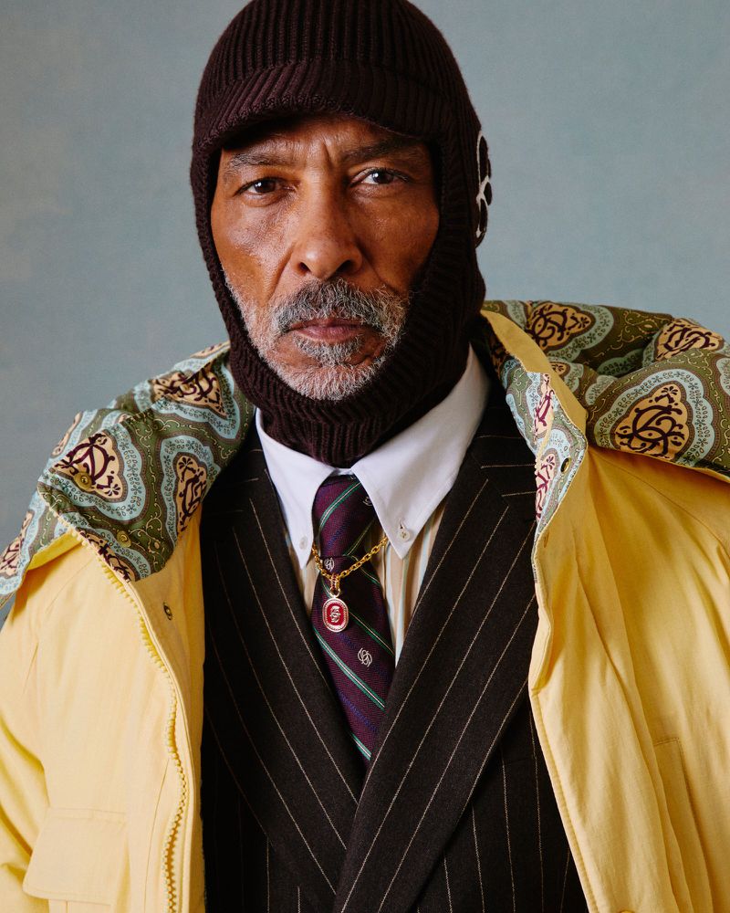Mature African American man in pinstripe suit and brown knit balaclava, layered with a yellow jacket; studio shot on light-blue background.