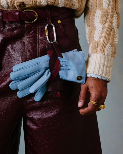 A stylish person wears maroon leather pants with a braided belt, a beige cable-knit sweater, and carries light blue leather gloves.