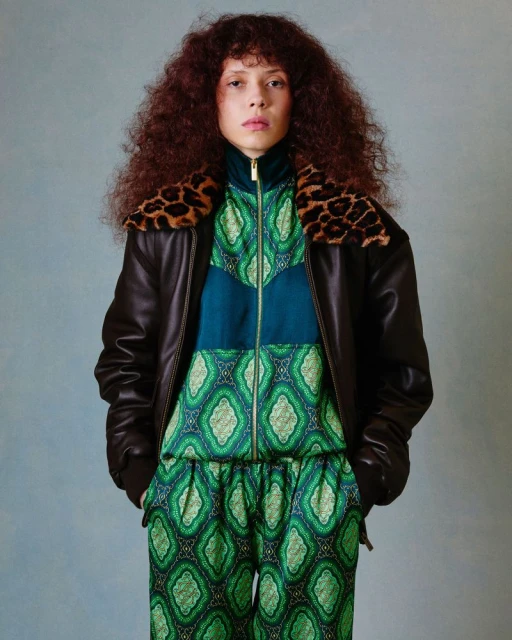 A woman with curly hair in a green patterned jumpsuit and brown leather jacket with a leopard print collar poses in a studio.