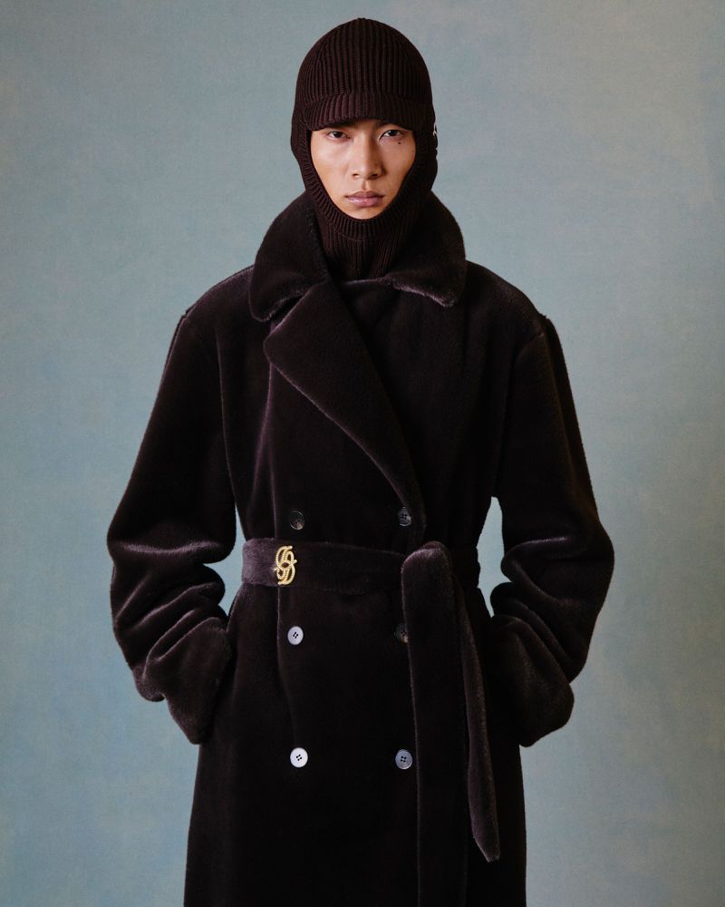 East Asian male model wearing a dark brown belted velvet coat and knit visor balaclava against a muted textured backdrop.