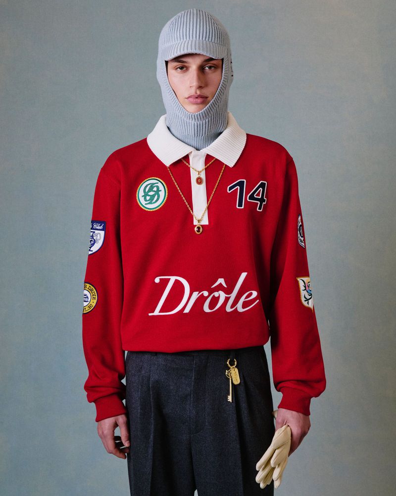 A model poses indoors wearing a red, long-sleeve polo shirt, grey pants, and a grey knitted balaclava with attached visor and gold necklace.