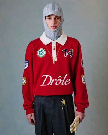 A model poses indoors wearing a red, long-sleeve polo shirt, grey pants, and a grey knitted balaclava with attached visor and gold necklace.