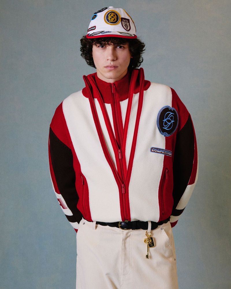A young man models a color-blocked track jacket with a baseball cap indoors. He wears light-colored pants and a black belt with a key charm.