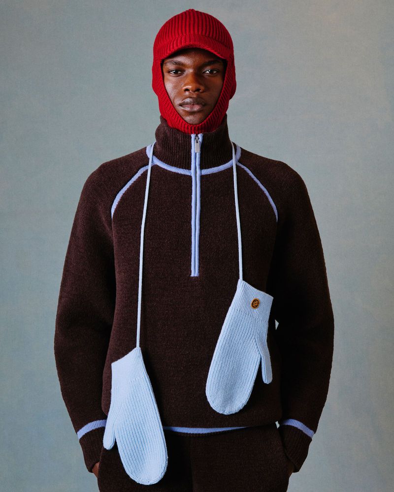 Male model wearing a brown knit sweater and matching pants, with a red knit hat and light blue mittens on strings. Fashion shot.