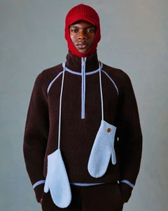 Model wearing a red balaclava, brown quarter-zip sweater with blue piping, and hanging blue mittens for Drole De Monsieur