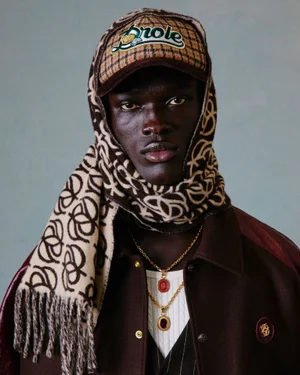 Close-up portrait of a Black man wearing a Drôle de Monsieur cap, patterned headscarf, and layered gold pendant necklaces.
