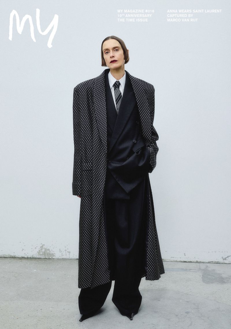 Model poses in oversized suit and coat for a fashion magazine cover, wearing dark makeup and a tie in a minimalist studio.