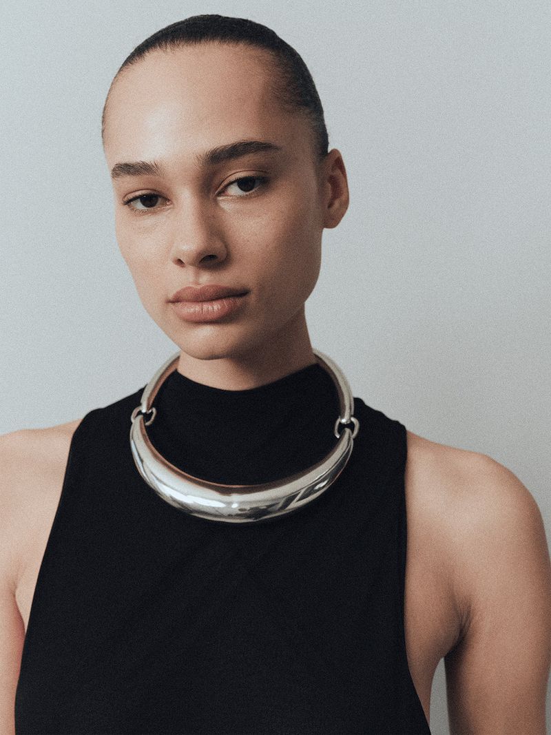 Close-up studio shot of a woman wearing a silver choker necklace and black top against a grey backdrop.