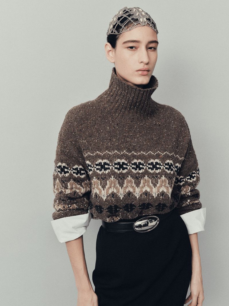 Portrait of a young woman wearing a brown patterned sweater, black skirt, and jeweled cap. Fashion editorial shot in a studio.