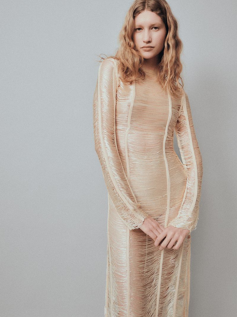 A model poses in a textured cream fringe long-sleeve dress against a neutral grey studio backdrop.