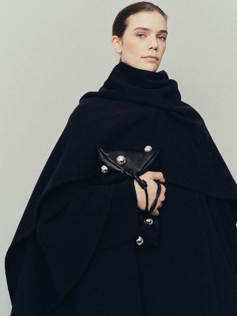 A woman in a dark wool shawl and coat stands in front of a light grey background, holding a small dark leather purse.