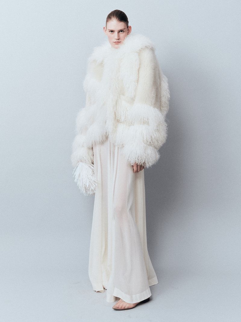 A person poses in a full-length shot, wearing a white, feathered jacket and a long, flowing white dress against a light grey background.