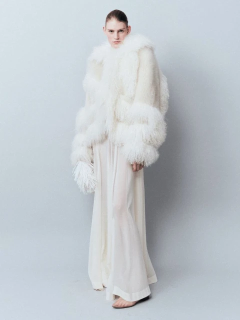 A person poses in a full-length shot, wearing a white, feathered jacket and a long, flowing white dress against a light grey background.