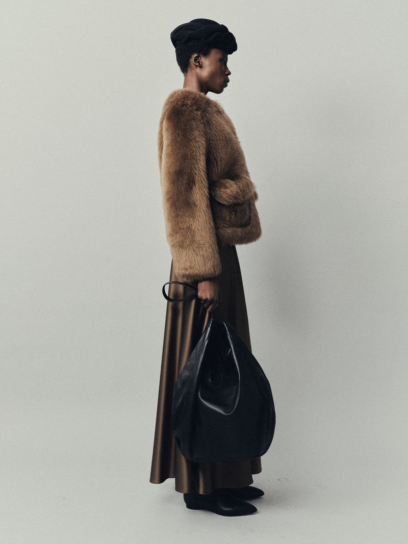 Elegant woman in a fur jacket and full skirt carrying a large black leather bag poses in a studio with a neutral background.