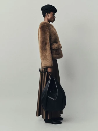 Elegant woman in a fur jacket and full skirt carrying a large black leather bag poses in a studio with a neutral background.
