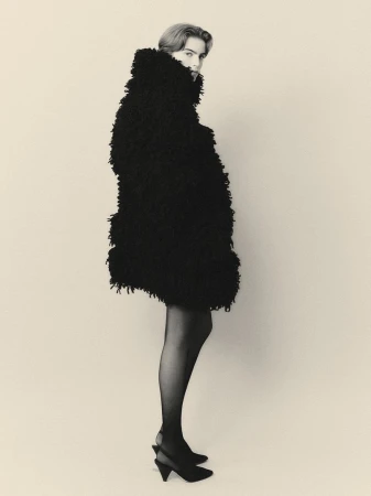 Full-length shot of a woman in black coat, black tights, and black slingback heels, posing in a studio with a neutral background.