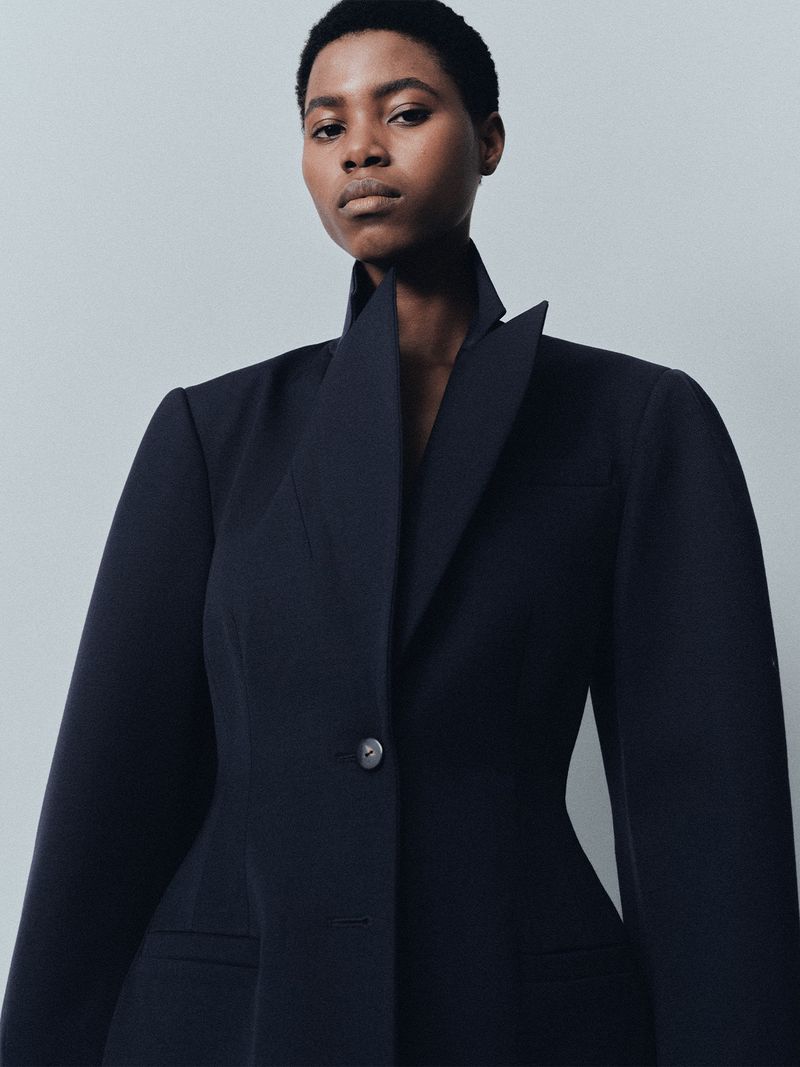 Close-up studio shot of a Black woman in a tailored black blazer with a wide, angled collar, looking directly at the camera.