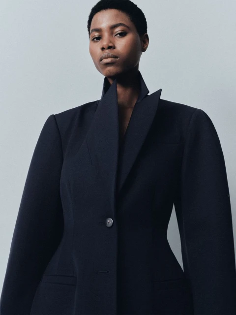 Close-up studio shot of a Black woman in a tailored black blazer with a wide, angled collar, looking directly at the camera.