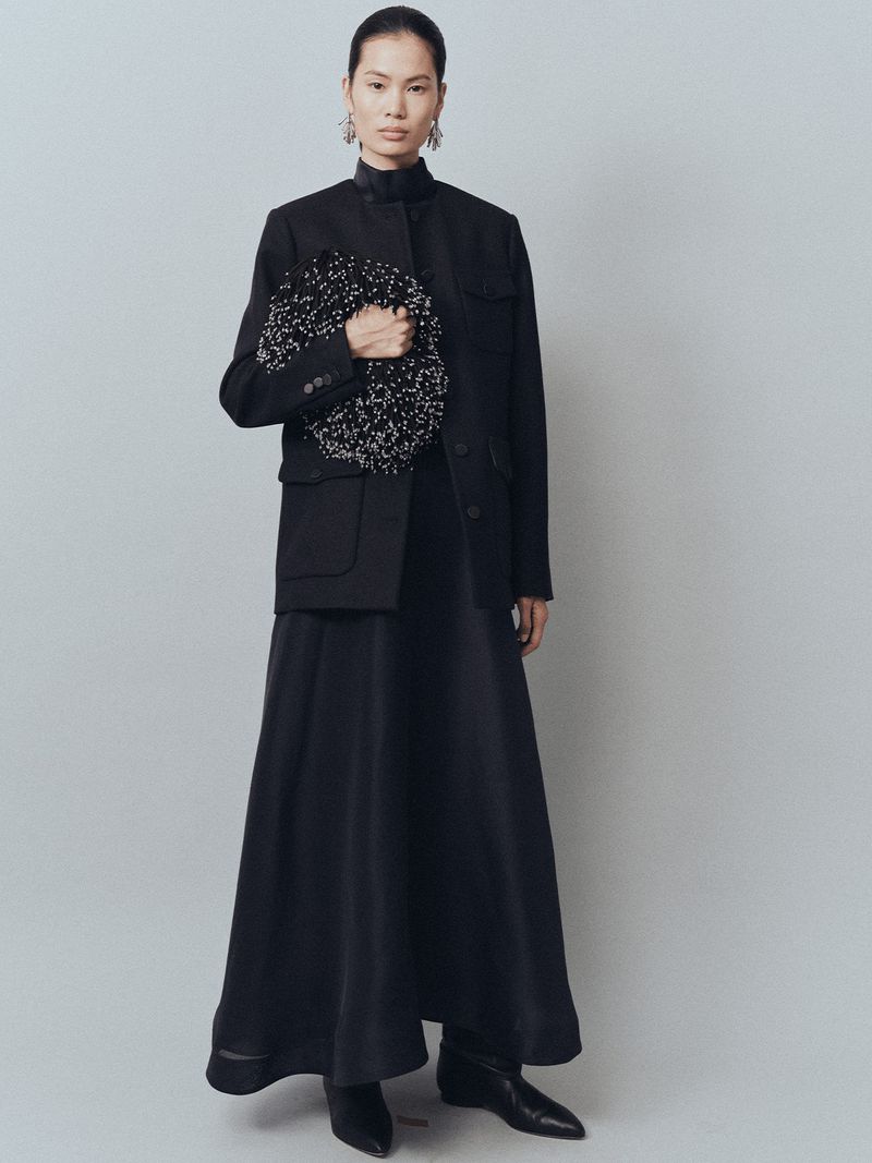 Elegant Asian woman in a black skirt suit holding a beaded bag, posing in a brightly lit photo studio against a light gray backdrop.