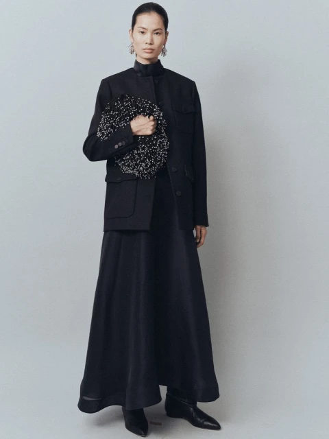Elegant Asian woman in a black skirt suit holding a beaded bag, posing in a brightly lit photo studio against a light gray backdrop.