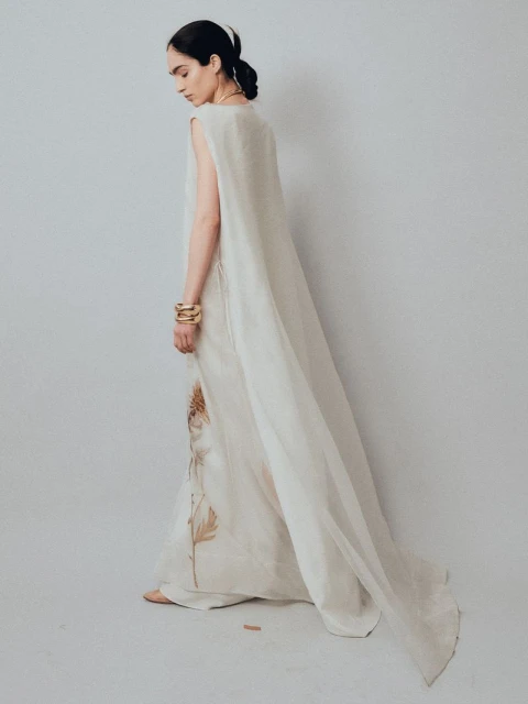 An adult woman in an elegant ivory dress stands in a neutral studio, accented by gold bracelets and stylized feather details on the dress.