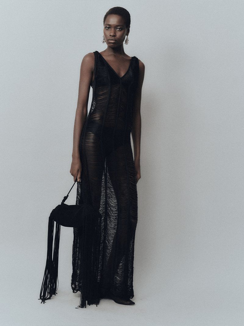 A black woman poses in a sheer black gown with a matching black fringed purse against a neutral studio backdrop.