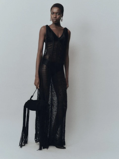 A black woman poses in a sheer black gown with a matching black fringed purse against a neutral studio backdrop.