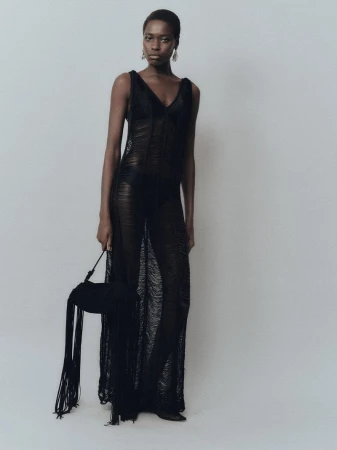 A black woman poses in a sheer black gown with a matching black fringed purse against a neutral studio backdrop.